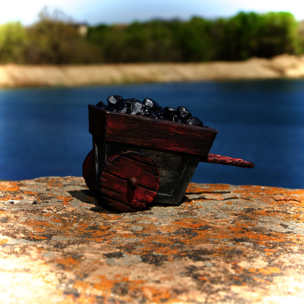 The Goblin Mine Cart, resting alone on a rock ledge overlooking a lake. The tiny cart is painted dot look like hammered metal, with heavy wood trim, wheels, and handles. The cart is filled with black pebbles that look like coal.