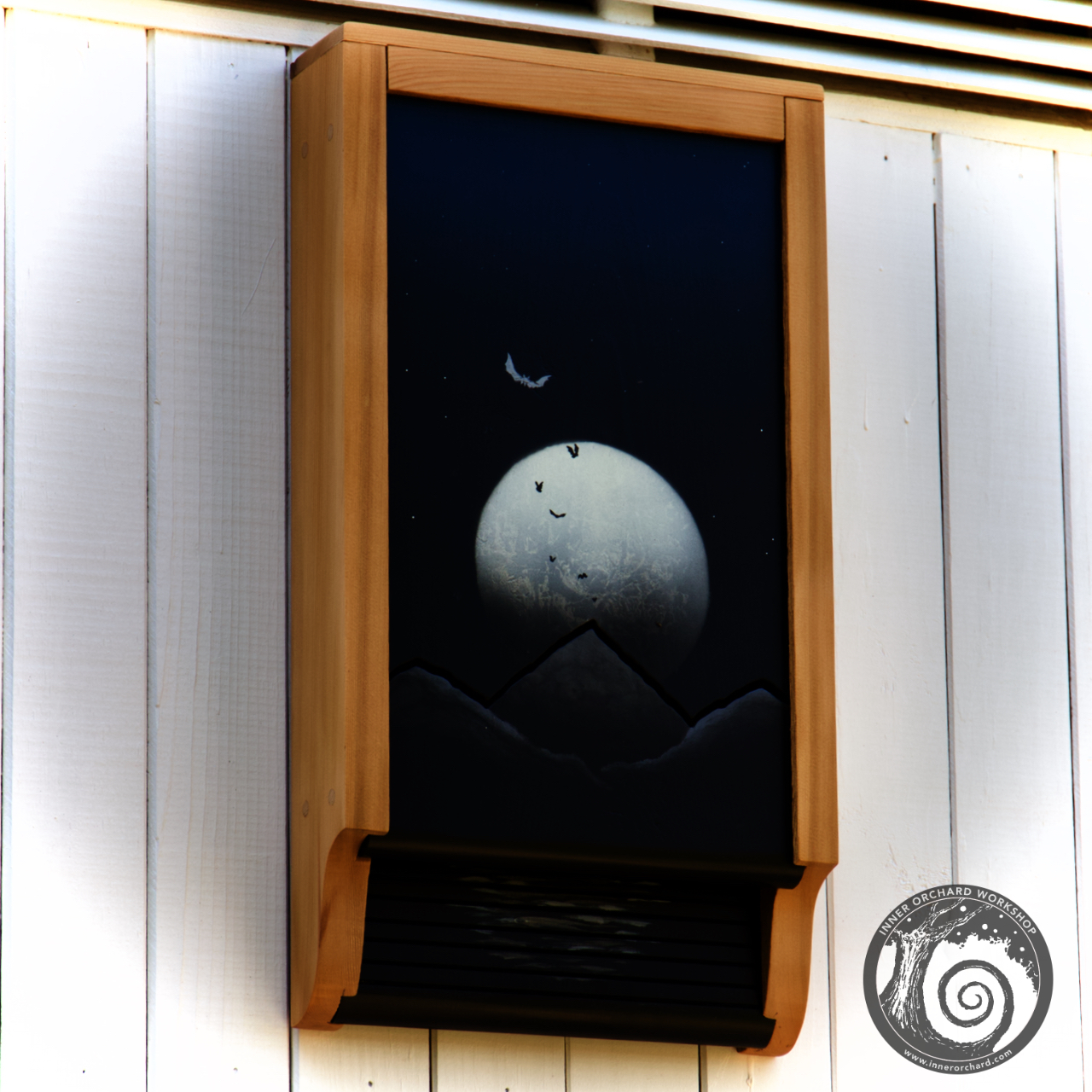 Image shows Moonrise Bat house mounted on the side of a white building. The wooden framework surrounds the image of a night sky with a beautiful moon rising over the mountains. The silhouettes of several bats stand out against the moon.